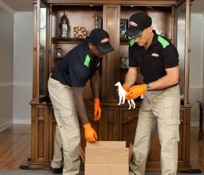 Two SERVPRO employees packing contents from a china cabinet in a living room.
